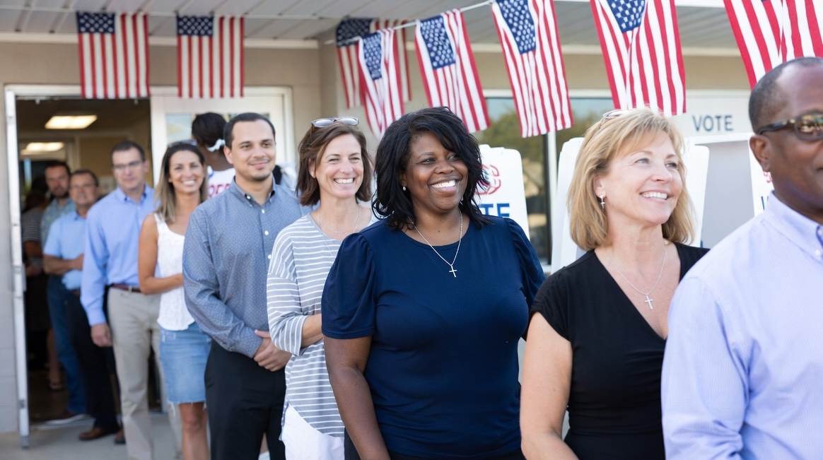 A young Christian woman with a cross necklace holds her ballot at a voting booth, with American flags in the background